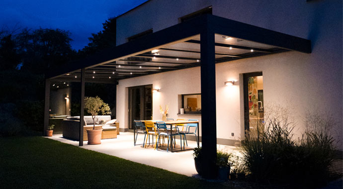 Moderne, anthrazitfarbene Terrassenüberdachung bei Nacht mit integrierten LED-Spots, die den Essbereich und die Lounge-Ecke auf der Terrasse hell ausleuchten.