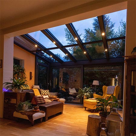 Stimmungsvolle LED-Beleuchtung im Wintergarten Gemütlich eingerichteter Wintergarten bei Abenddämmerung mit warmweißen LED-Spots, die harmonisch in die Dachsparren der Glaskonstruktion integriert sind.